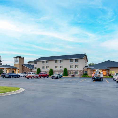 A building resembling a hotel with a parking lot, several vehicles parked, and a clear, blue sky with thin clouds in the background.