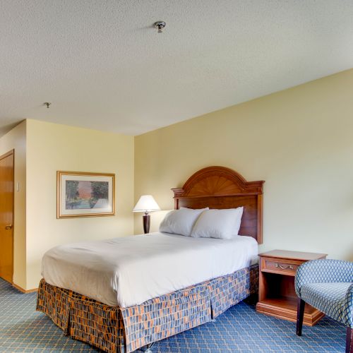 This image shows a hotel room with a bed, two nightstands, a chair, a lamp, and a framed picture on the wall, with a door and hallway visible.