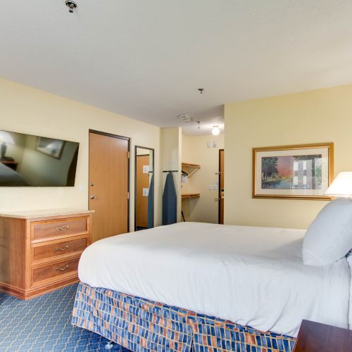 A hotel room with a bed, TV, dresser, microwave, mini-fridge, ironing board, and wall art, featuring yellow walls and carpeted flooring.