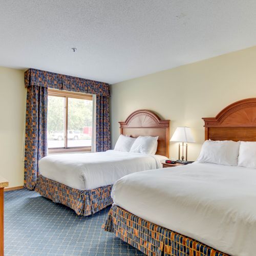 The image shows a hotel room with two double beds, a window, a desk, and a chair. The decor features patterned curtains and bedskirts.