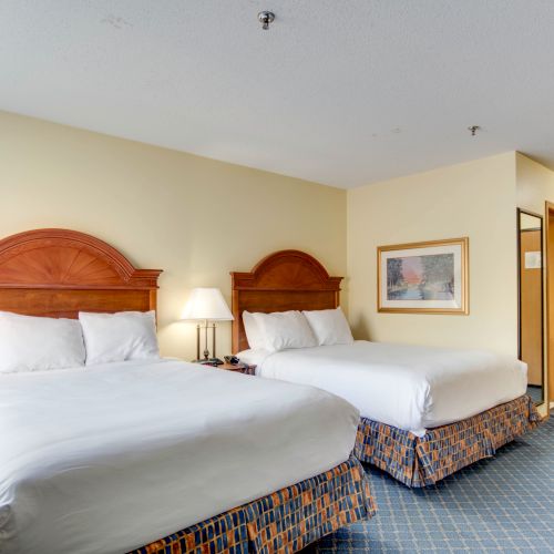 The image shows a hotel room with two double beds, a bedside lamp, a framed picture on the wall, and a bathroom door in the background.