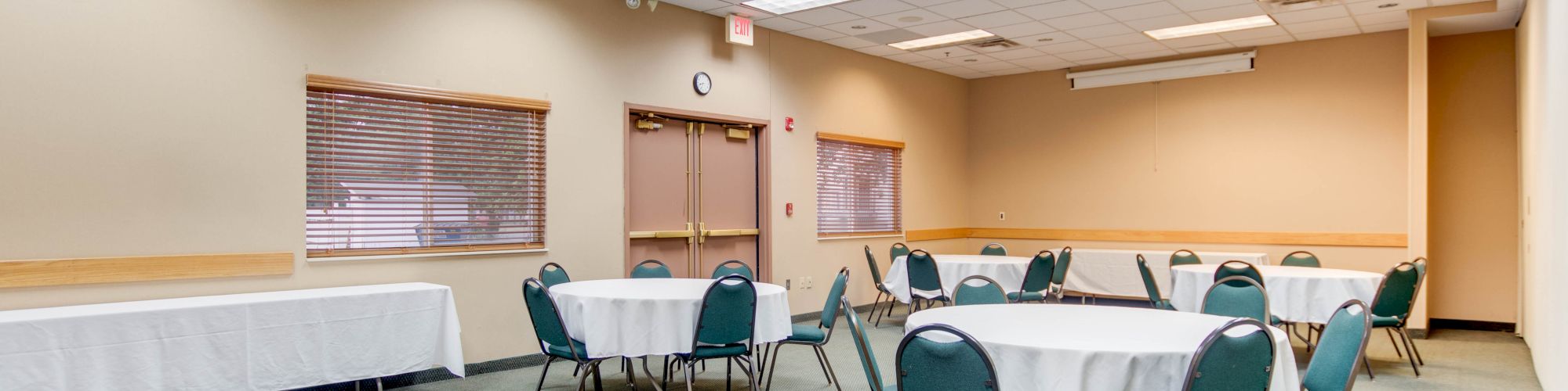 A small conference room with round tables and green chairs, white tablecloths, a side table, fluorescent lighting, and a carpeted floor.