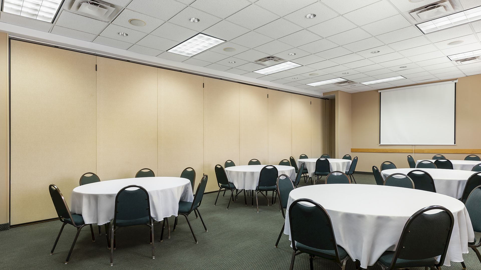 A conference or event room with round tables draped in white cloth, black chairs, and a projector screen at the front, set up for a meeting or banquet.
