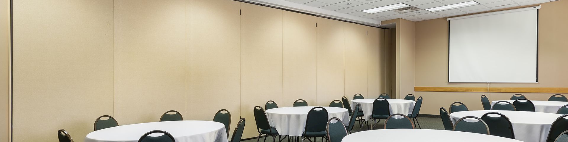 A conference or event room with round tables draped in white cloth, black chairs, and a projector screen at the front, set up for a meeting or banquet.