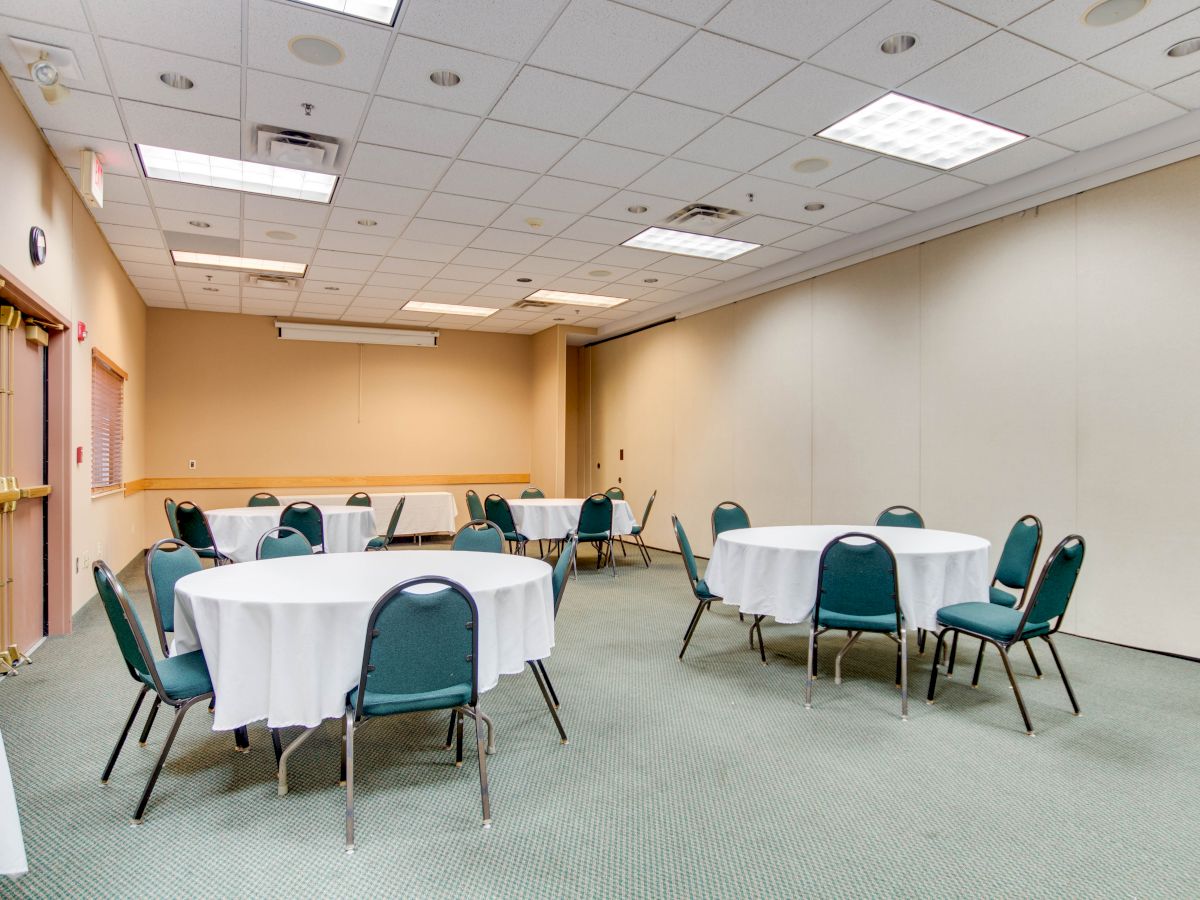 A conference room with round tables covered in white tablecloths and green chairs arranged around them. The room is well-lit with ceiling lights.