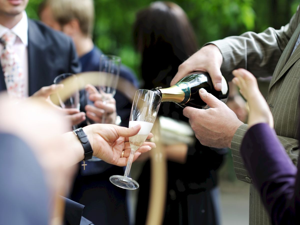 People are gathered, dressed formally, while a person pours champagne into a glass, suggesting a celebratory event or gathering.