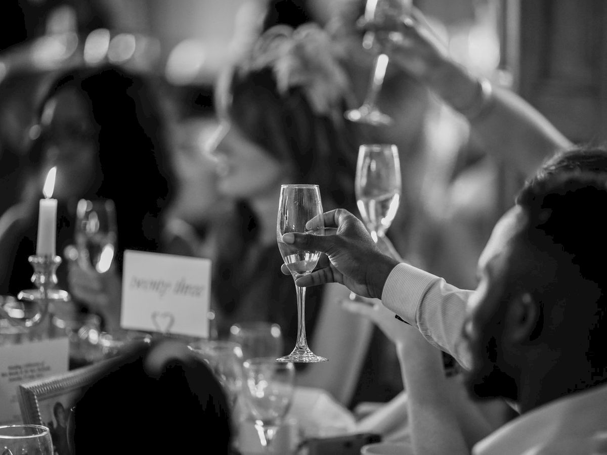 People are raising wine glasses in a toast at a gathering or celebration. A lit candle and table decorations are visible.