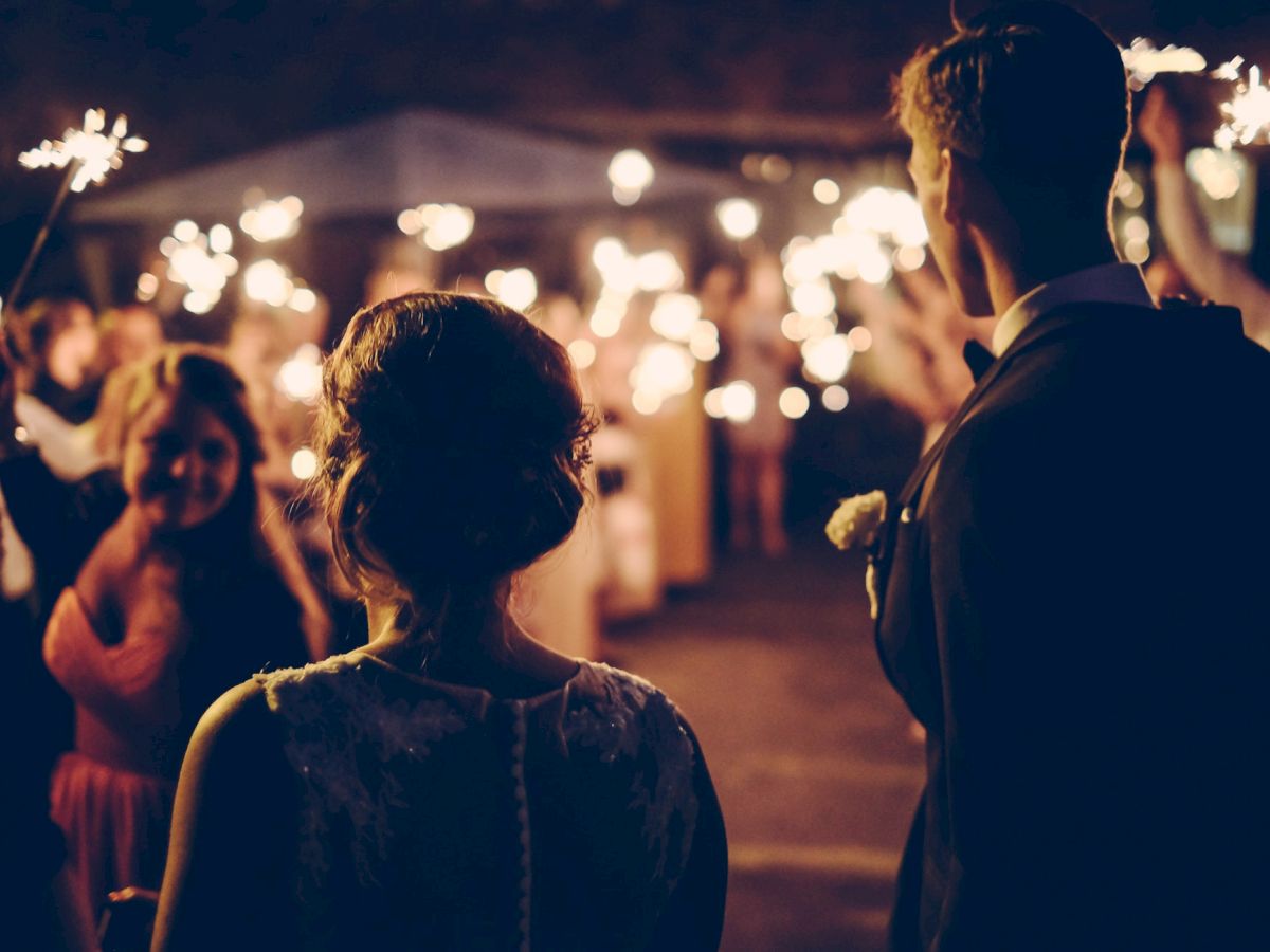 A couple stands before a crowd holding sparklers in a festive, warmly-lit celebration, possibly a wedding or party, at nighttime.