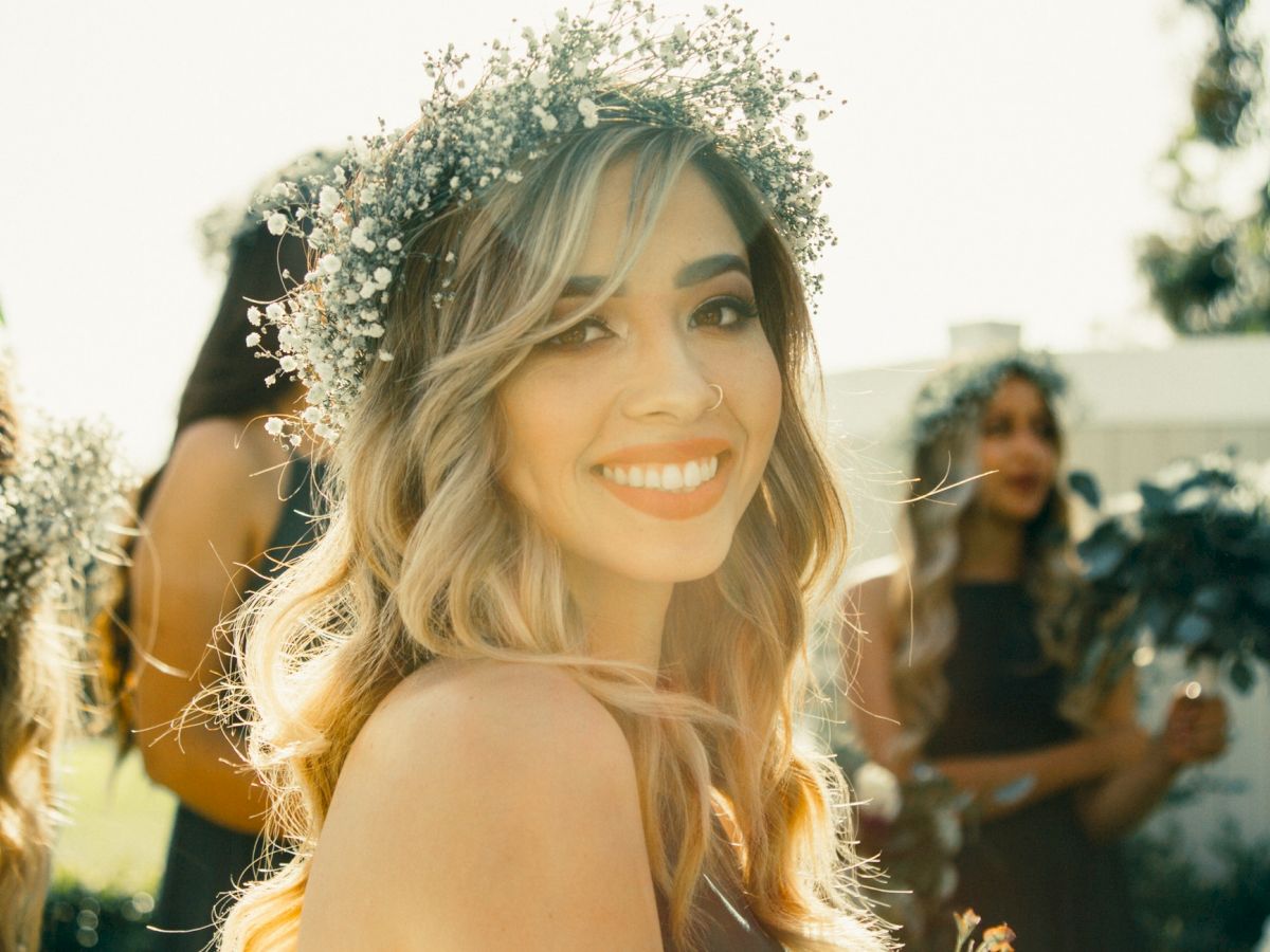 A woman with a floral crown smiles at the camera, surrounded by other women who also have floral crowns, outdoors in a sunny setting.