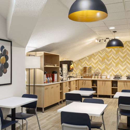 A bright, modern break room with light wood cabinets, small tables and blue chairs, zigzag yellow wall, and pendant lights.