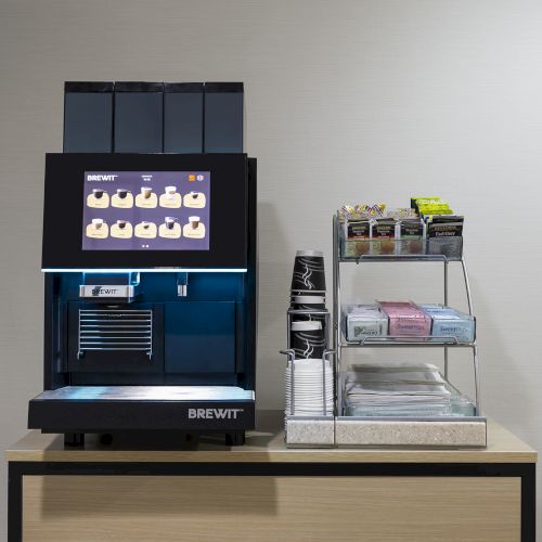 A black coffee machine on a wooden table beside a metal rack with cups, napkins, and lids in a minimal caf&eacute; setup.