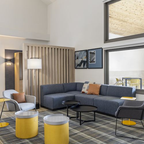 A modern living room with a gray sectional sofa, yellow accent stools, a black coffee table, and a light wood screen separating a cozy seating area, large windows.