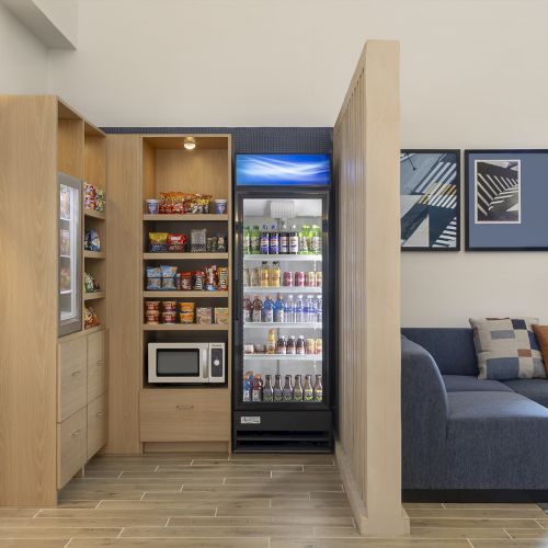 A small kitchenette area with wooden cabinets, a microwave, shelves of snacks, and a tall beverage fridge next to a blue sofa in a bright lounge.