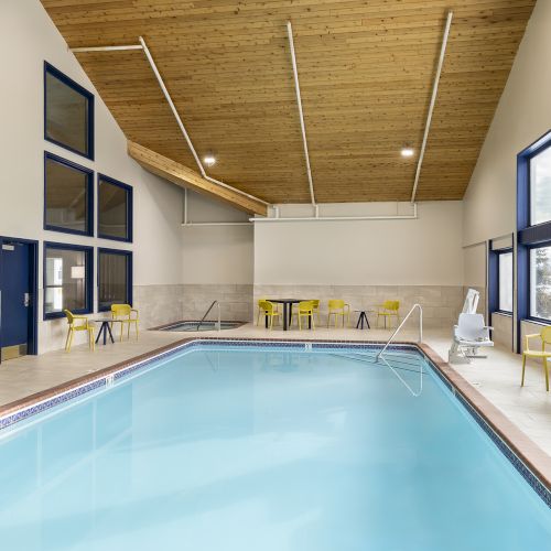 Indoor swimming pool with clear blue water, sunny windows, a wooden ceiling, tiled surround, and yellow chairs along the walls.