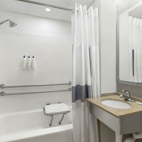 A modern accessible bathroom with a walk-in tub, grab bars, a shower head, a foldable seat, and a granite countertop vanity with a mirror.