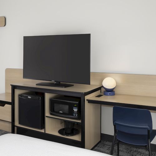 A modern hotel desk setup with a mounted TV, mini-fridge, microwave, and storage cubbies; a blue chair at a long wooden console desk.