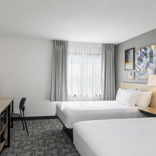 A modern hotel room with two double beds, a flat-screen TV on a dresser, a small desk and chair, a window with curtains, and framed art on the wall.