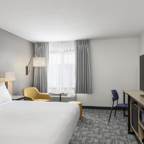 A modern hotel room with a large bed, seating area, desk, TV, and window with gray curtains, in neutral tones.