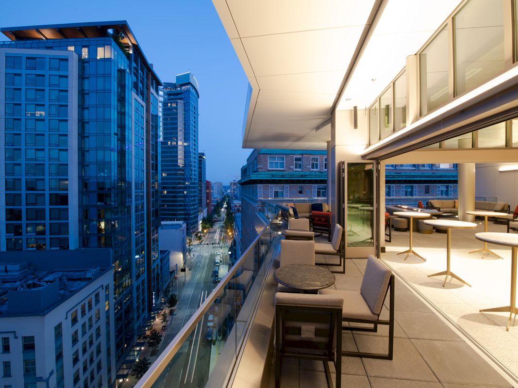 A modern cityscape at dusk with high-rise buildings and a stylish outdoor patio area featuring tables and chairs on a balcony.