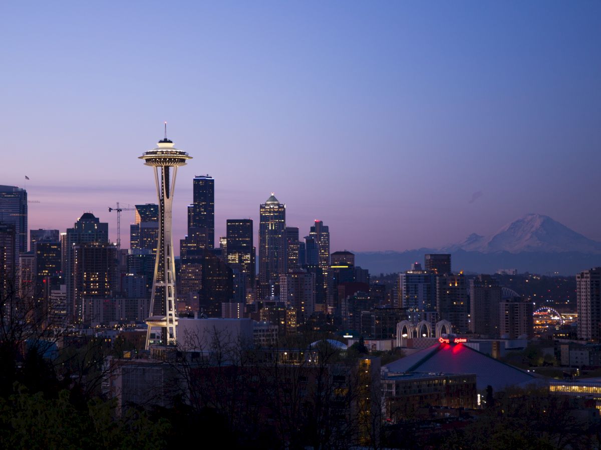 The image features the Seattle skyline at dusk, showcasing the Space Needle and Mount Rainier in the background, with the city lights starting to illuminate.