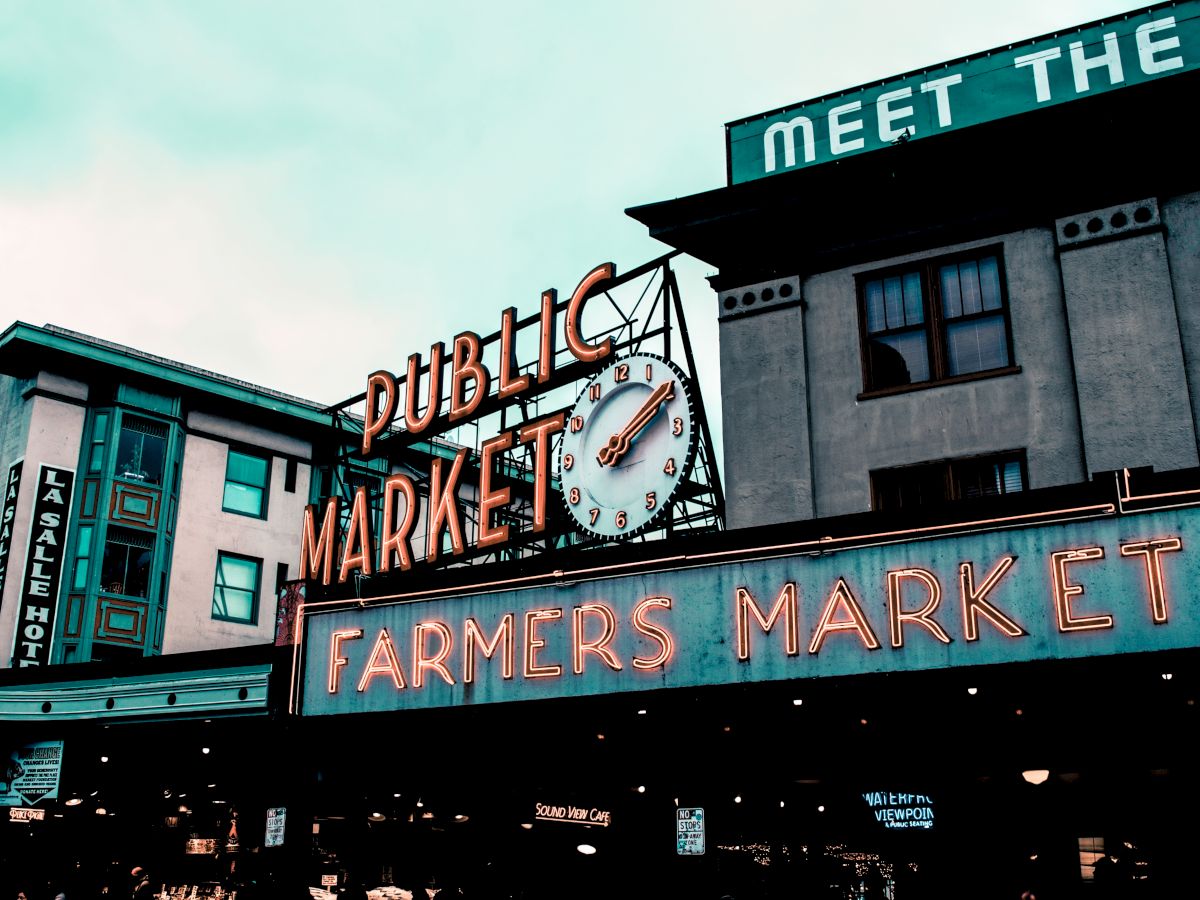 The image shows the iconic entrance sign of a public farmers market, with neon lights and a clock, surrounded by various buildings and shops.