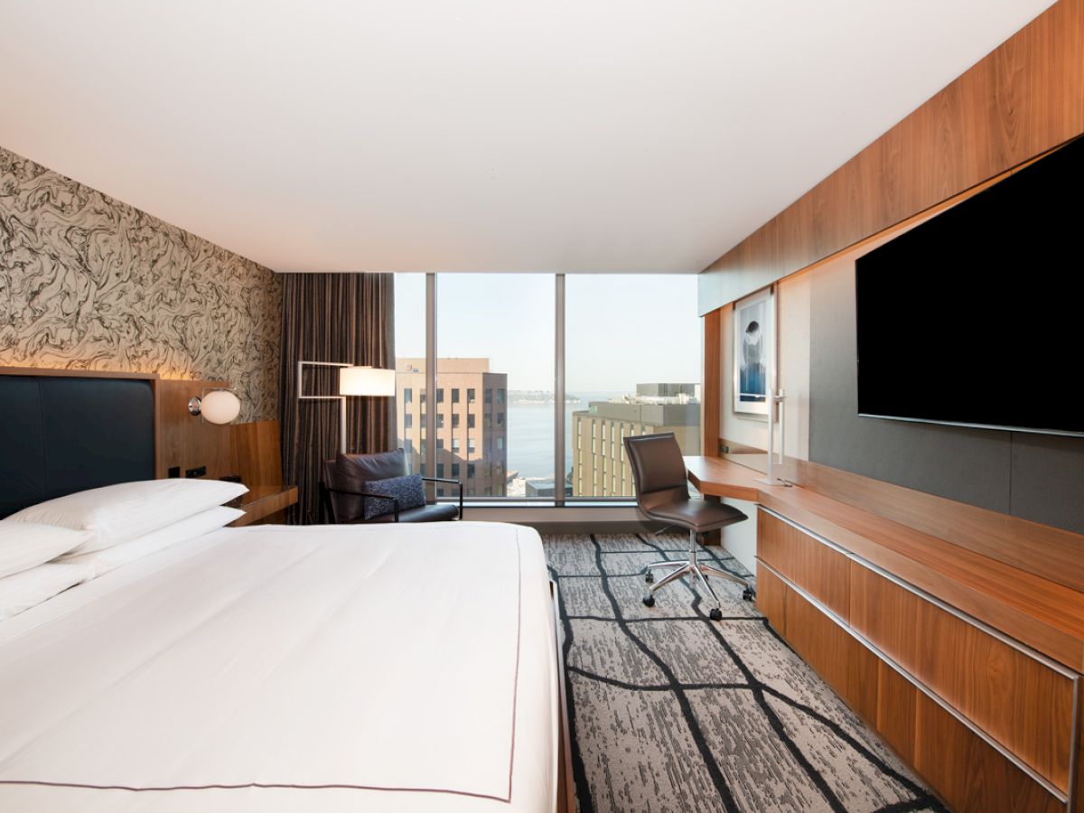 This image shows a modern hotel room with a large bed, a desk and chair, a flat-screen TV, and a view of city buildings through a large window.