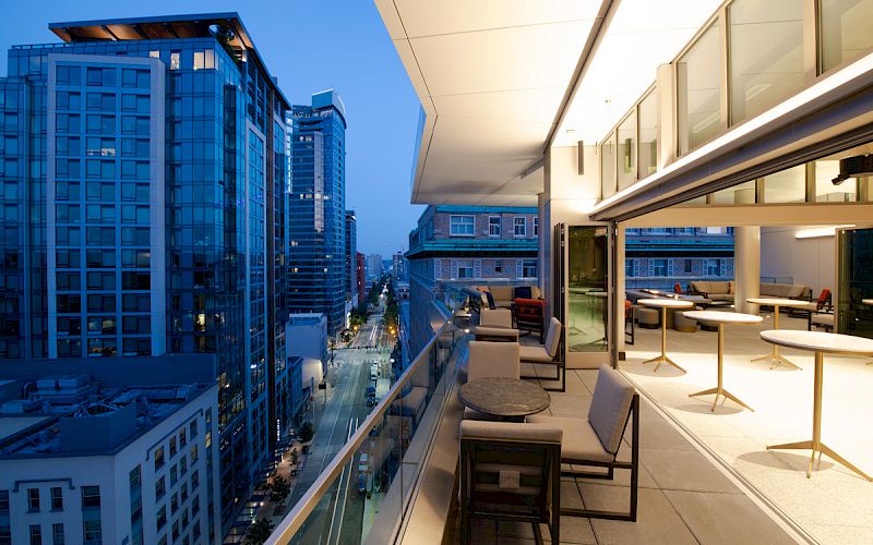 An evening cityscape shot showing a modern balcony with tables and chairs overlooking a street flanked by tall buildings.